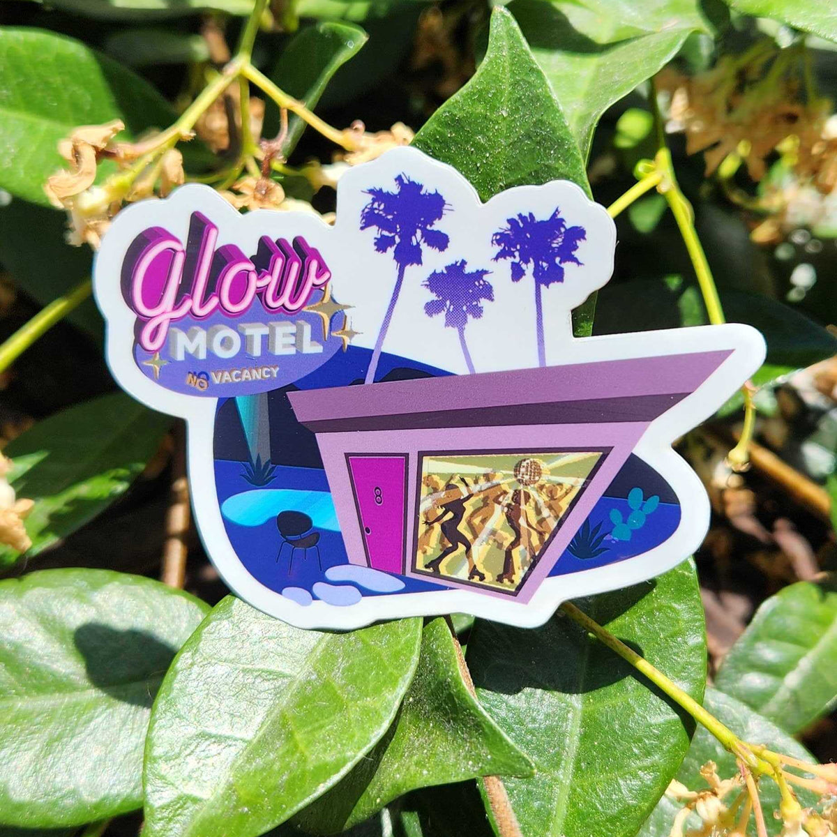 A die-cut sticker featuring a retro motel scene with the text 'Glow Motel 24 Vacancy,' purple palm trees, and a motel building, photographed outdoors against green leaves.