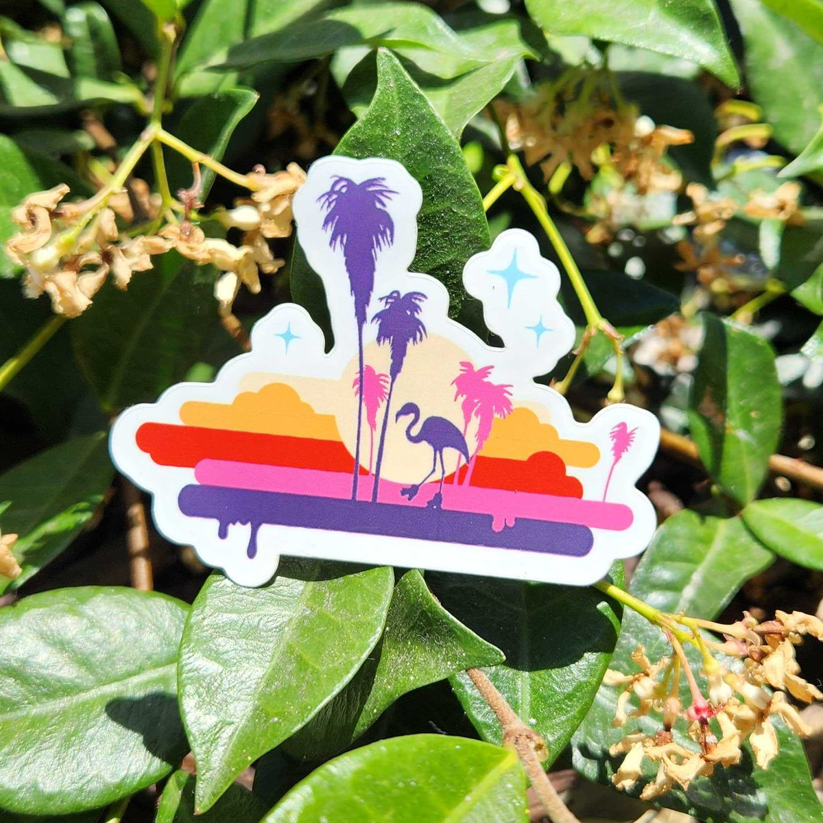 A die-cut sticker featuring a retro sunset beach scene with palm trees, a flamingo, and colorful horizontal stripes, shown placed on green leaves outdoors.