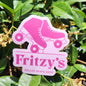 A die-cut sticker featuring a pink roller skate graphic and the text 'Fritzy's Roller Skate Shop,' photographed outdoors against green leaves and small flowers.