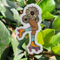 A die-cut sticker featuring an illustrated figure with flower heads and roller skates, placed on green leaves outdoors.