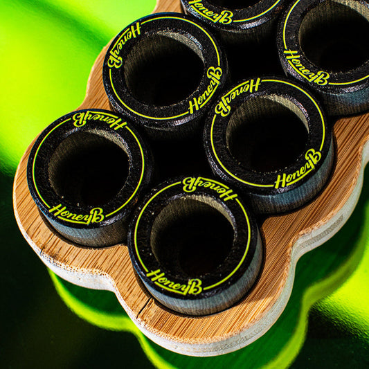 Six black shot glasses with neon yellow 'HoneyB' branding, arranged in a wavy bamboo holder against a bright green background.