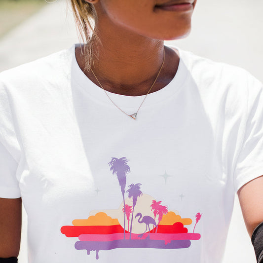 A white short-sleeve t-shirt with a colorful graphic of palm trees, flamingos, and sunset stripes in purple, pink, orange, and red. 