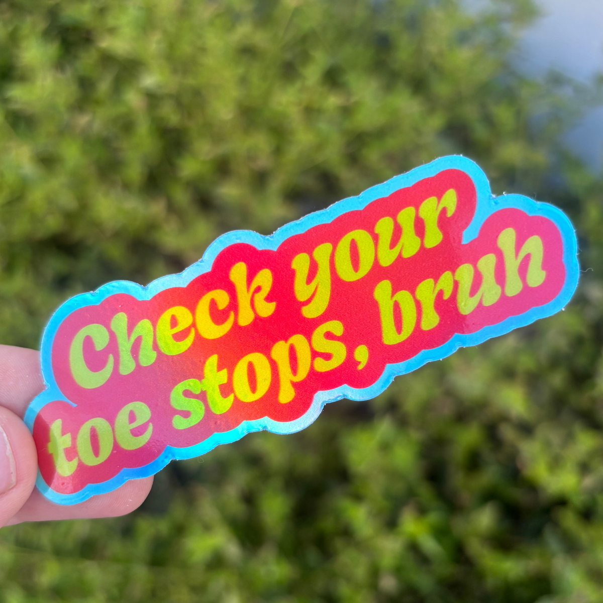A hand holds a die-cut sticker with the text 'Check your toe stops, bruh' in neon yellow-green lettering on a red background with a blue outline, against a blurred green outdoor background.