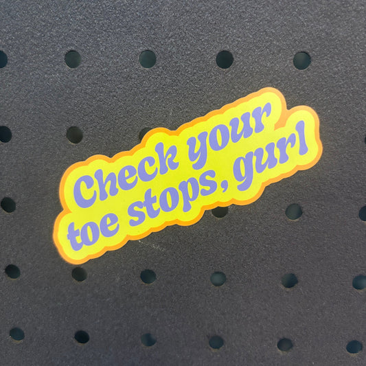 A die-cut sticker with the phrase 'Check your toe stops, gurl' in purple text outlined in yellow, placed on a dark perforated foam background.